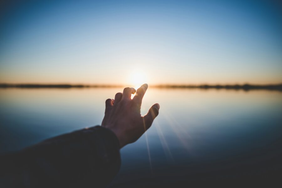 Hand reaching towards the rising sun over a calm body of water, symbolizing freedom from guilt, hope, and the opportunity for a new beginning filled with peace and purpose.