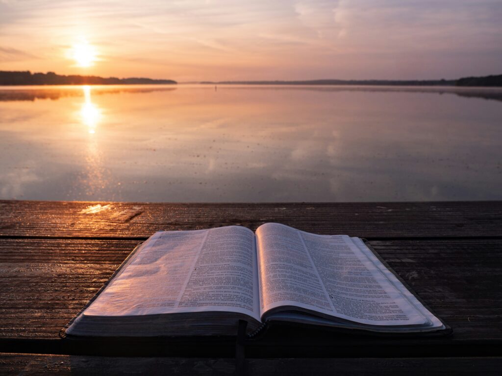 Open Bible on a wooden surface by the water at sunrise, symbolizing quiet moments for hearing God's voice, prayer, and spiritual reflection.