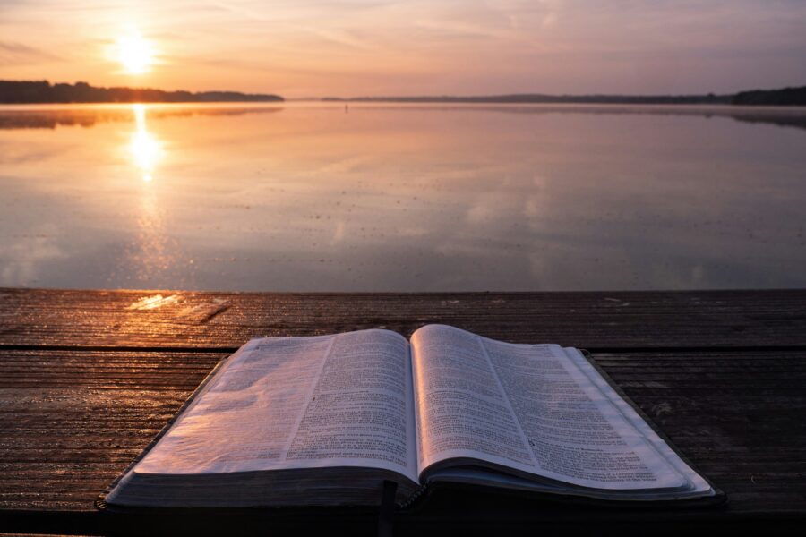 Open Bible on a wooden surface by the water at sunrise, symbolizing quiet moments for hearing God's voice, prayer, and spiritual reflection.