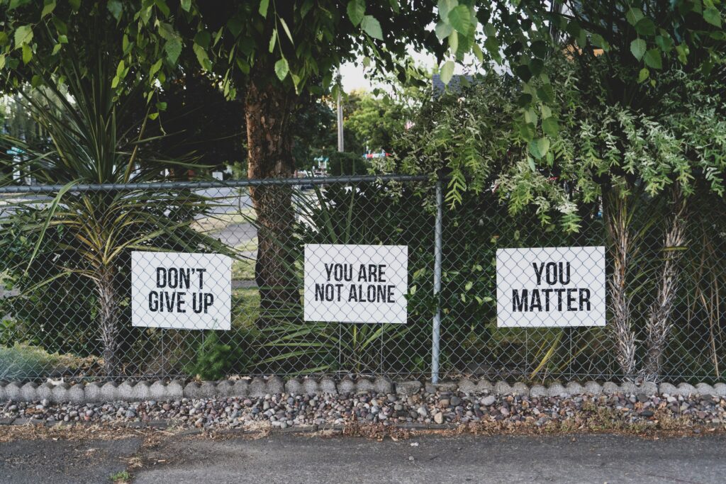 Three signs on a fence offer encouragement: 'Don't Give Up,' 'You Are Not Alone,' and 'You Matter.' A visual reminder to release worry, trust in God, and find peace in the small, meaningful ways He shows up in your life.