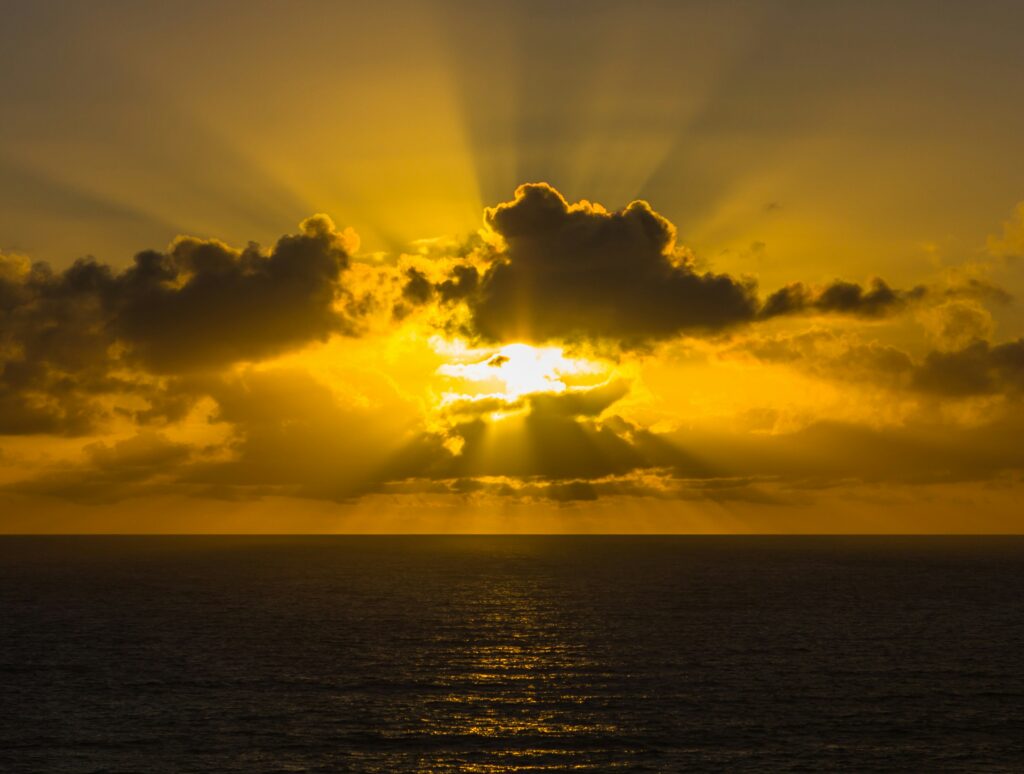 The sun shines through clouds over an endless ocean horizon at sunset, casting a golden glow on the water.