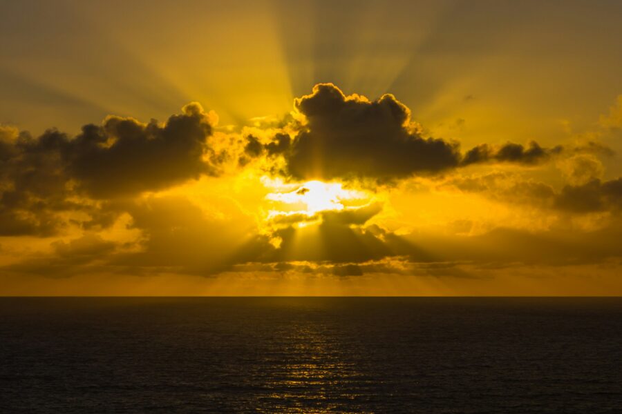 The sun shines through clouds over an endless ocean horizon at sunset, casting a golden glow on the water.