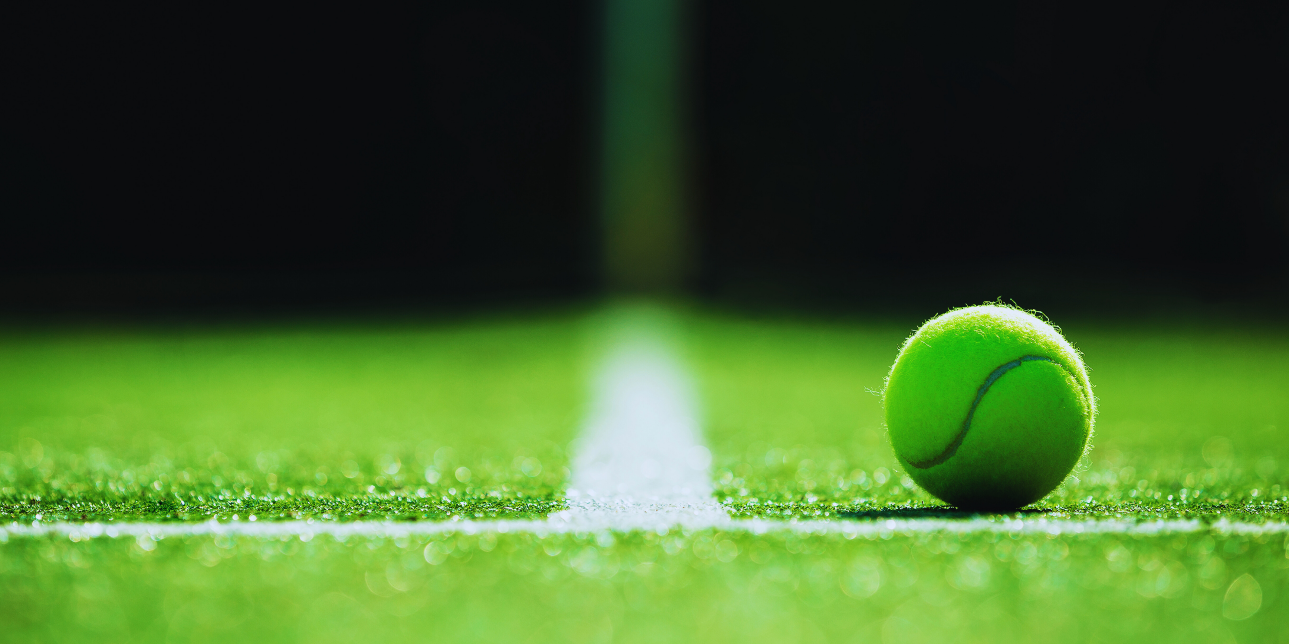 A close-up of a bright green tennis ball resting on a white line of a tennis court, with the background fading into darkness.