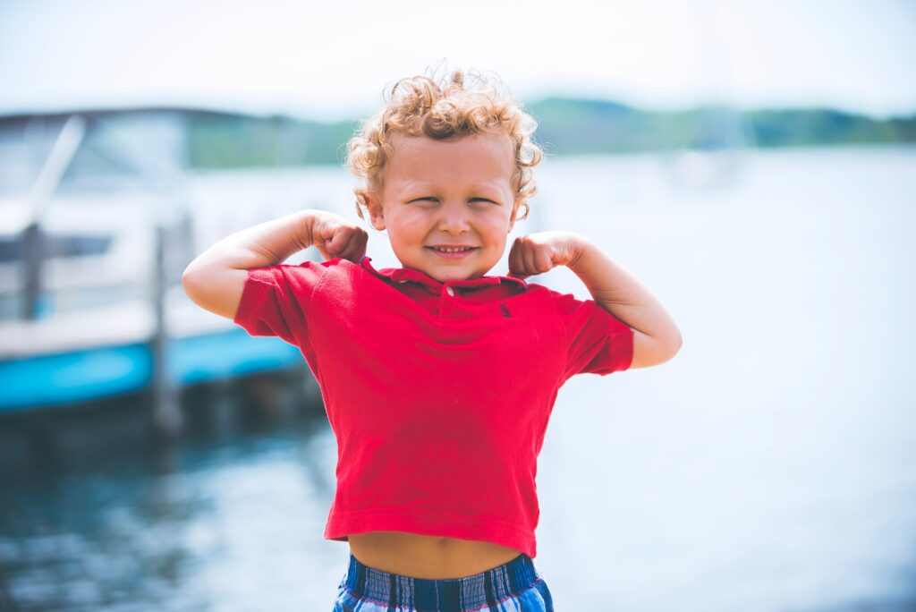 "A young child in a red shirt flexing their arms confidently, smiling at the camera with a bright background of water and boats."