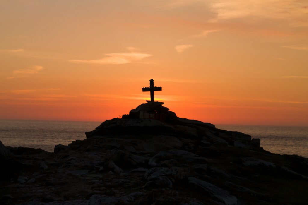 Silhouette of a cross on a rocky hilltop at sunset, overlooking the ocean, symbolizing the sacrificial love of Jesus.