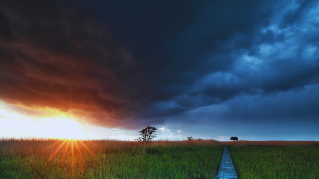 Dark storm clouds parting to reveal a bright sunrise over a grassy field with a wooden path leading forward.