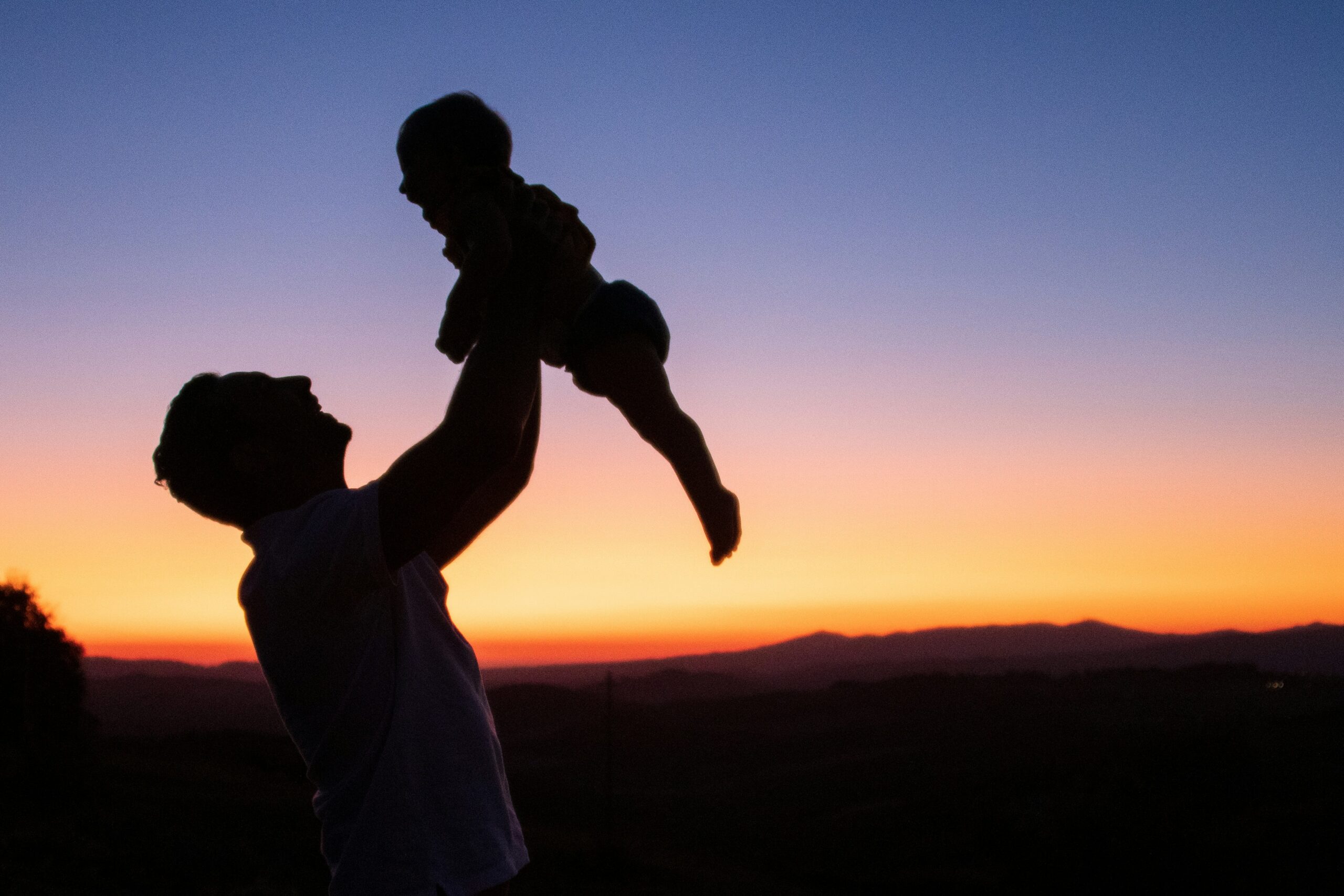 Silhouette of a father lifting his baby into the air at sunset with mountains in the background.